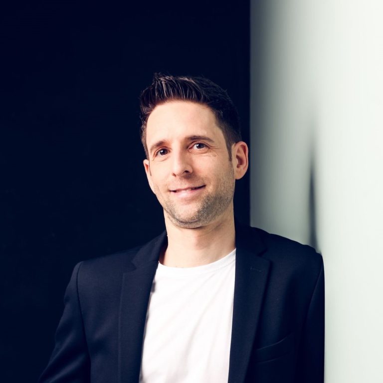 Mann in schwarzem Blazer und weißem T-Shirt, lächelt vor einem dunklen Hintergrund.
