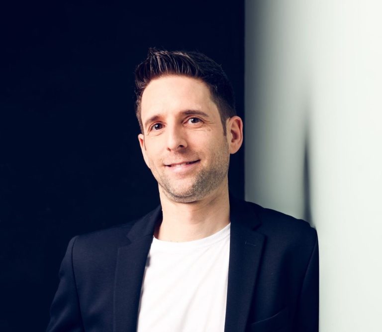 psychologischer-Berater-Coach-Mentor-Manager Mann in schwarzem Blazer, lächelt vor einem neutralen Hintergrund.