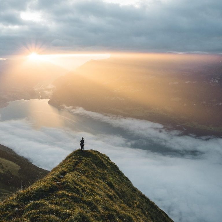 Person steht auf einem Berghang mit Blick auf einen Fluss und Sonnenstrahlen durch Wolken.