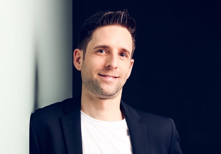 Mann in Blazer lehnt lässig an einer Wand und lächelt freundlich.