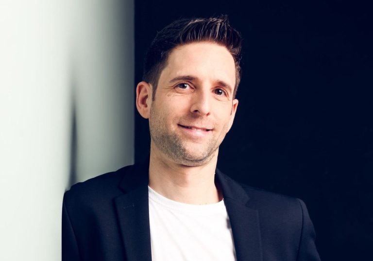 Mann in lässiger Kleidung, lächelnd vor dunklem Hintergrund, steht an einer Wand.