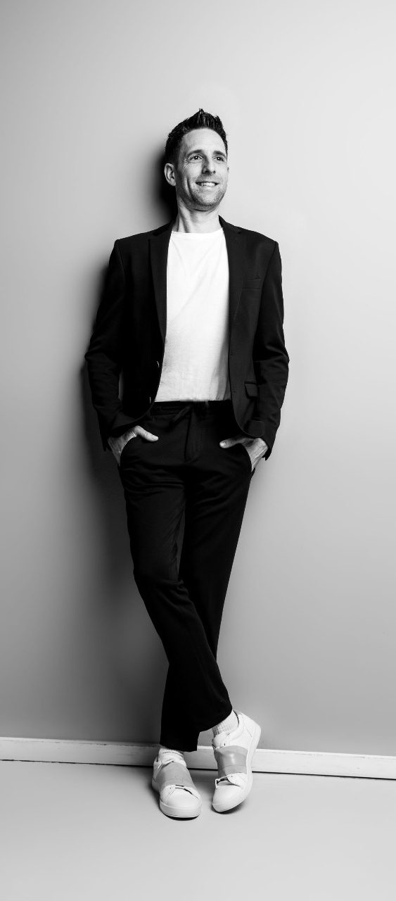 Person in Anzug und Sneakers lehnt an grauer Wand, lächelt leicht.